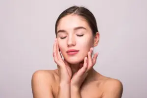 Against a plain light background, a young woman with bare shoulders gently touches her face and closes her eyes, reflecting on the refreshing effects of a hydrafacial at Refresh MedSpa in Cape Coral, FL.