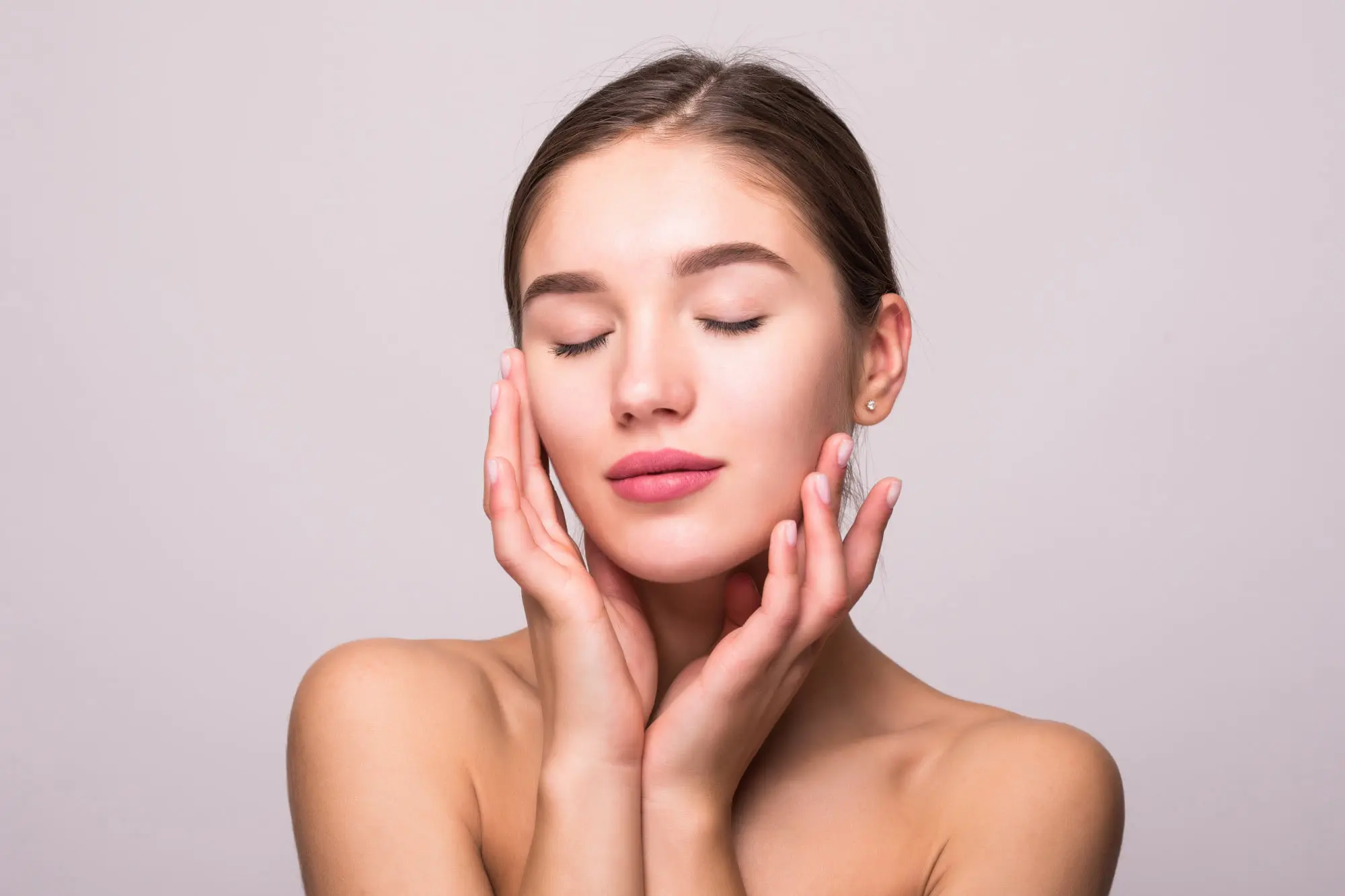 Against a plain light background, a young woman with bare shoulders gently touches her face and closes her eyes, reflecting on the refreshing effects of a hydrafacial at Refresh MedSpa in Cape Coral, FL.
