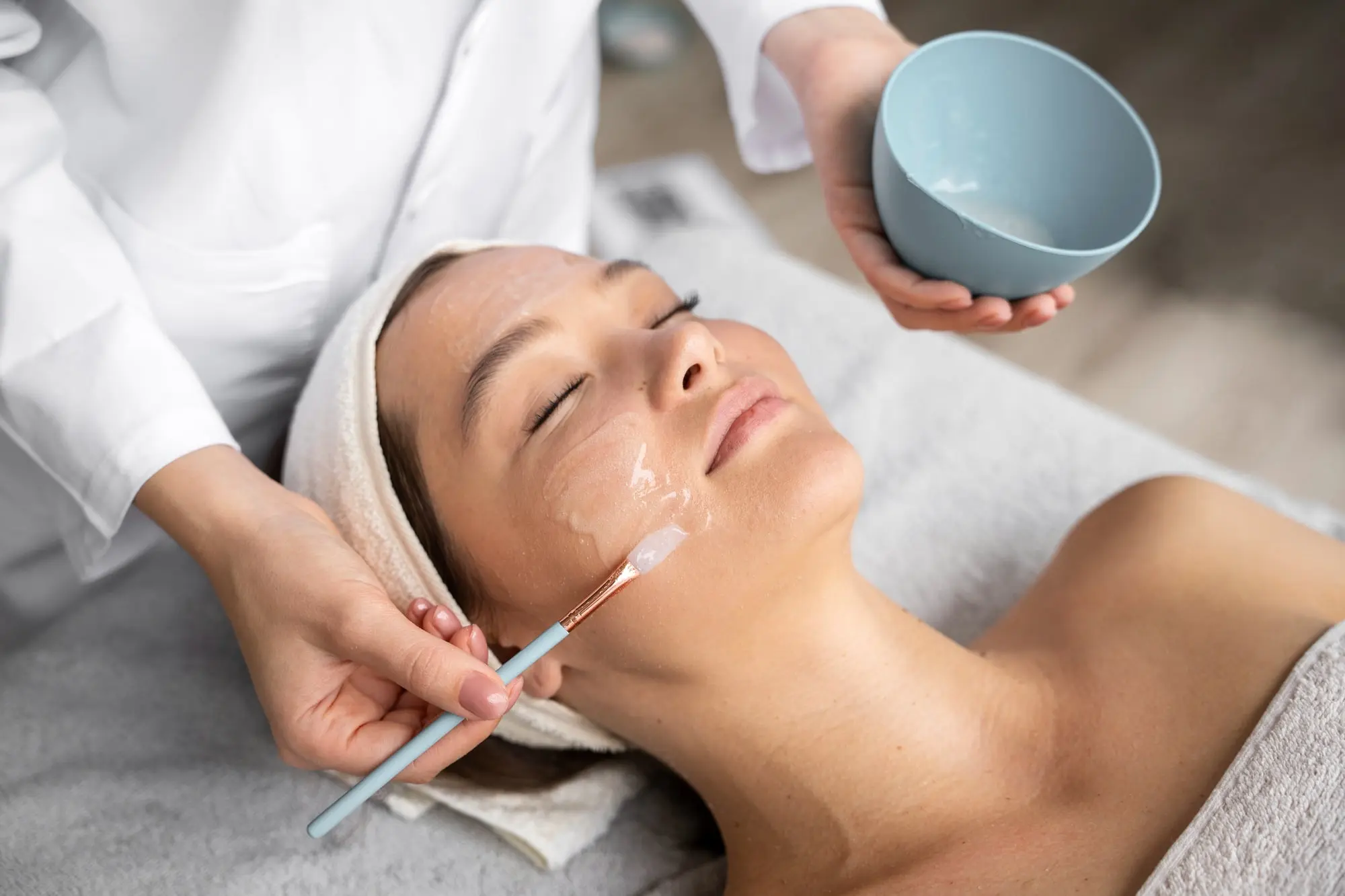 At Refresh MedSpa in Cape Coral, FL, a skincare specialist uses a brush to apply a facial mask from a bowl during a treatment—both hands are visible, suggesting you may wonder how often to get chemical peels.