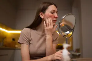In a kitchen, a woman with long brown hair checks redness on her cheek in a round mirror, thinking about rosacea flare-up triggers and common causes to avoid at Refresh MedSpa in Cape Coral, FL.