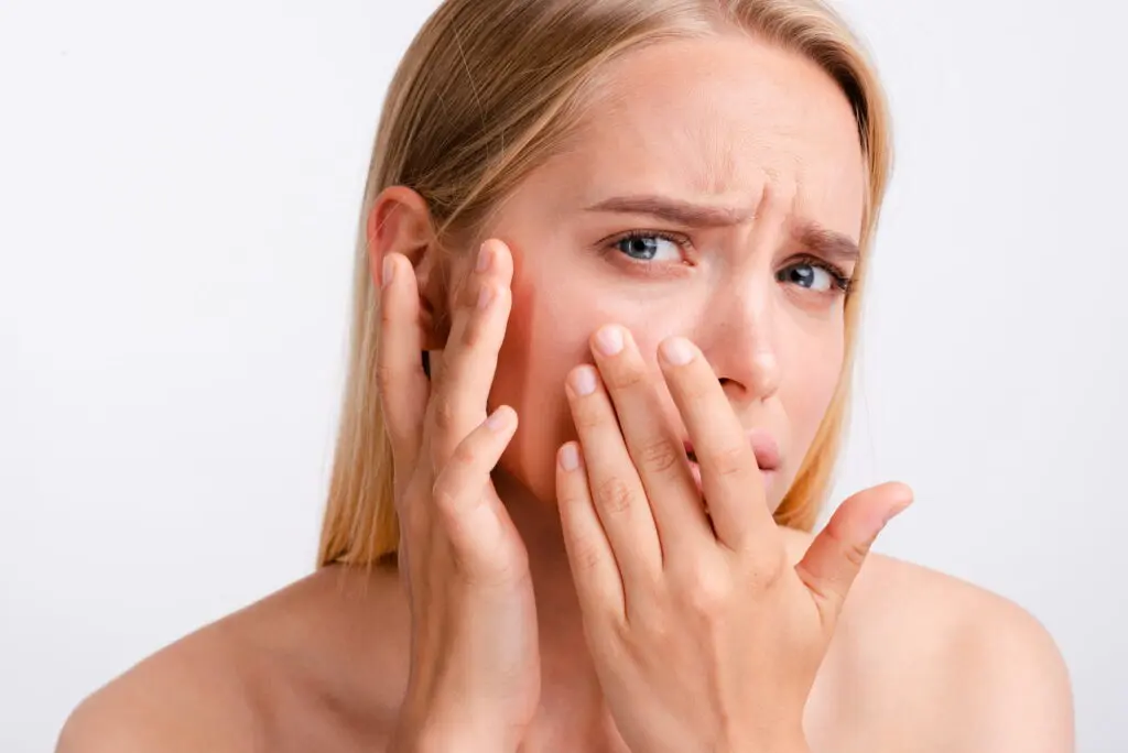 Against a plain background, a woman with long blonde hair looks concerned while touching her cheek and covering her mouth, appearing to contemplate the difference between dehydrated and dry skin at Refresh MedSpa in Cape Coral, FL.