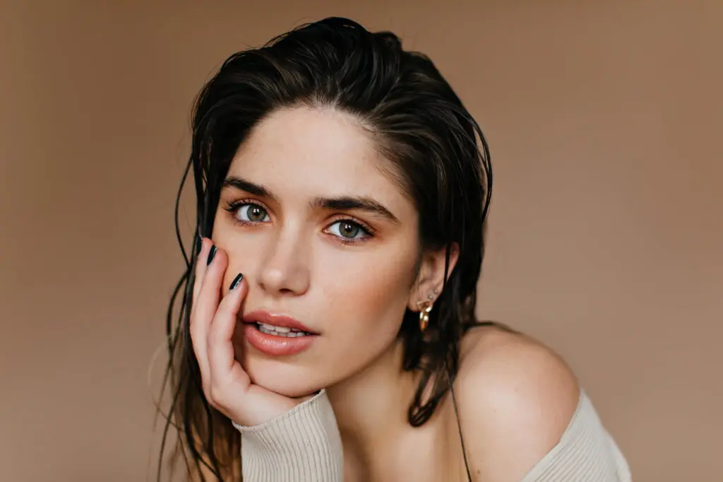 A woman with wet hair rests her face on her hand, gazing at the camera against a beige background, as if considering skincare options at Refresh MedSpa in Cape Coral, FL such as Hydrafacial vs Chemical Peel.