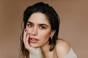 A woman with wet hair rests her face on her hand, gazing at the camera against a beige background, as if considering skincare options at Refresh MedSpa in Cape Coral, FL such as Hydrafacial vs Chemical Peel.