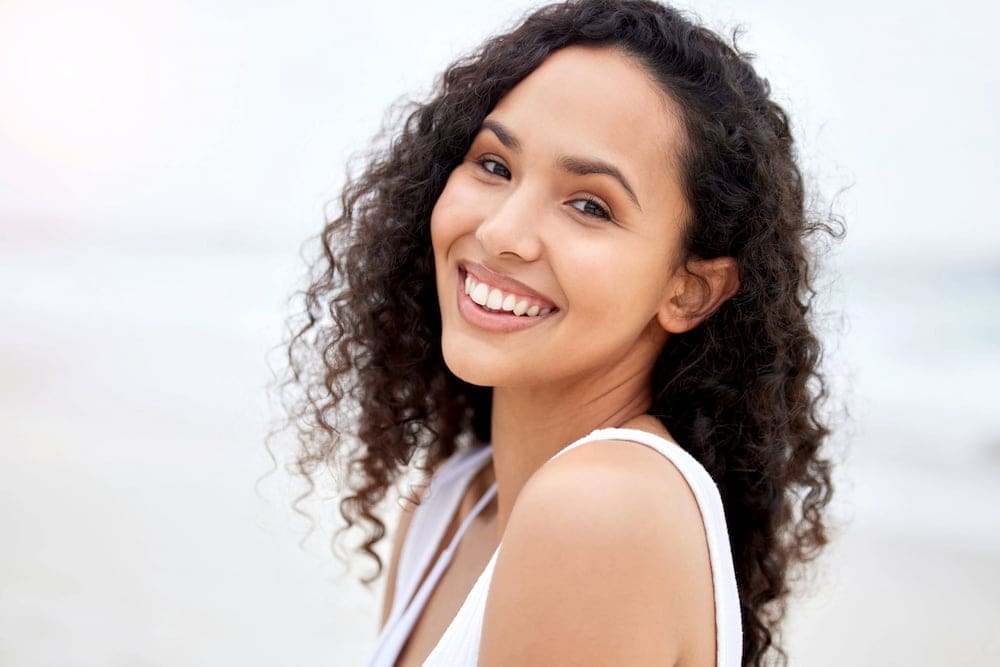 Smiling on the beach, a curly-haired person in a white top exudes confidence, possibly after Dermal Fillers at Refresh MedSpa.