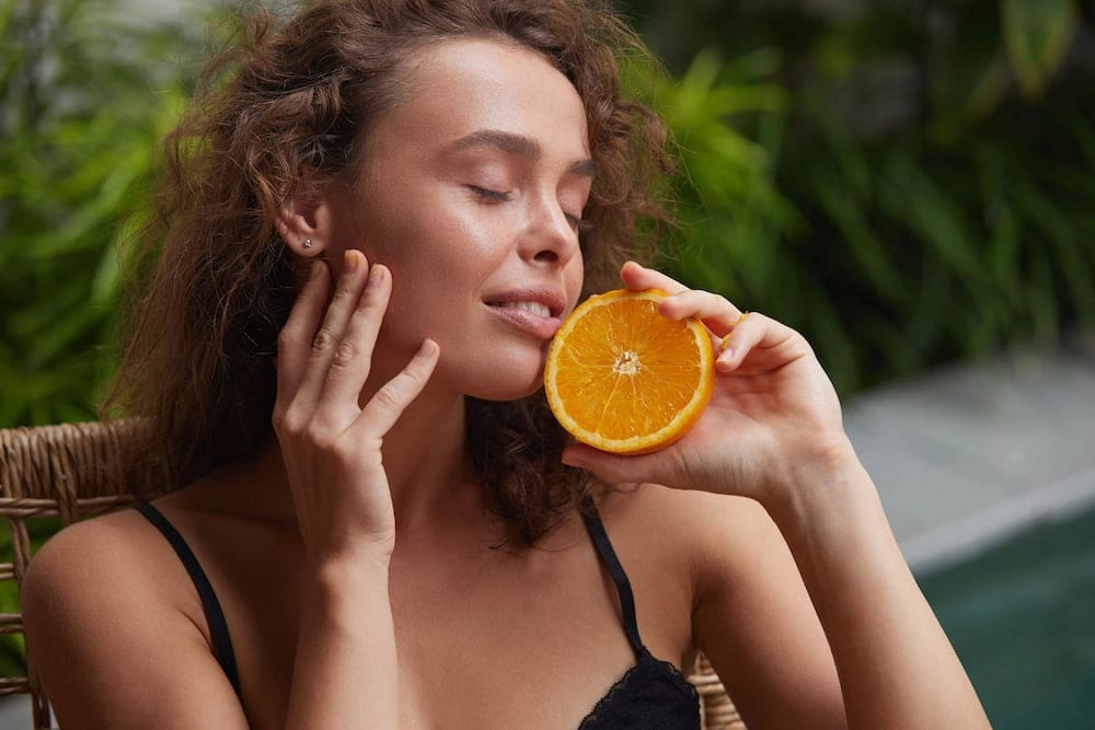 Amid green foliage, a woman with curly hair holds an orange near her face, glowing like after chemical peels at Refresh MedSpa.